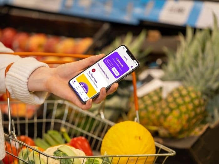 Woman carry a supermarket basket while holding a phone showing the Nectar app