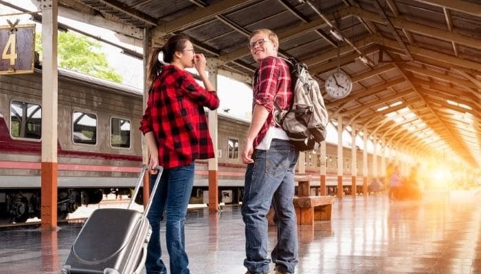Smiling young couple with luggage standing on a train platform with a train in the background.