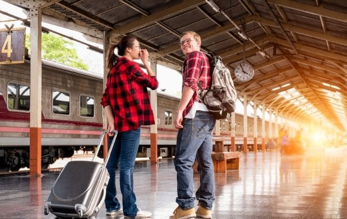 Smiling young couple with luggage standing on a train platform with a train in the background.