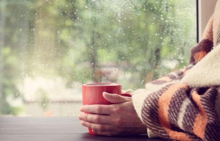 Person under a blanket holding a red cup in front of a rain covered window.