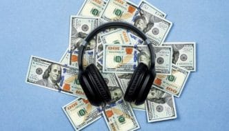 Black headphones on top of a pile of 100 dollar bills, placed on a plain blue background