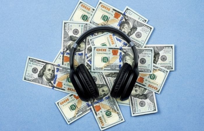 Black headphones on top of a pile of 100 dollar bills, placed on a plain blue background