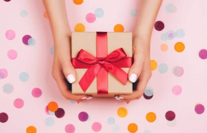 Hands holding a small present, with a red box, held over a pink background with confetti.