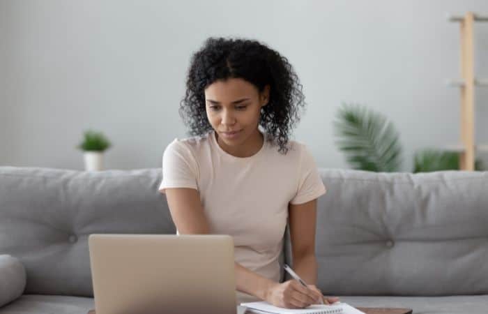 woman sitting on a sofa, looking at a laptop and writing notes