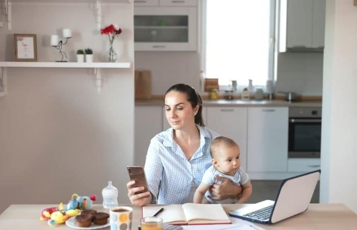 woman sitting at a kitchen table, holding a baby on one knee and looking at a phone in the other hand. There is a laptop and baby toys on the table.
