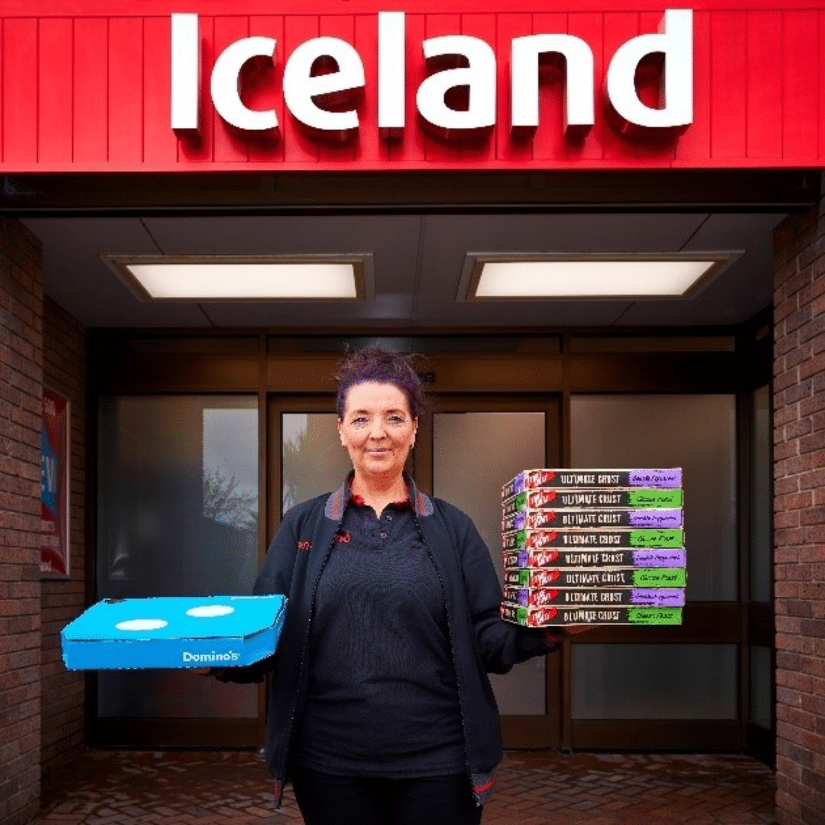 person standing outside an Iceland store. In their right hand, they are holding a Dominos pizza box. In their left hand, they are holding eight Iceland pizzas stacked up.