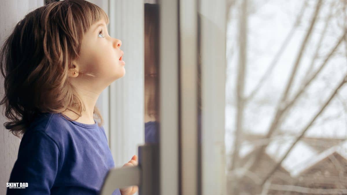 Child standing by a window at home in soft winter light, looking outside