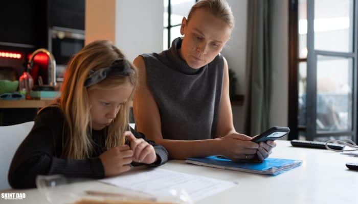 Parent and child sitting at a kitchen table, with the parent looking at a phone while the child works on paperwork