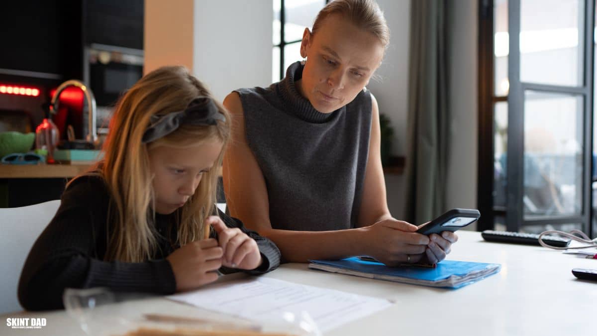 Parent and child sitting at a kitchen table, with the parent looking at a phone while the child works on paperwork