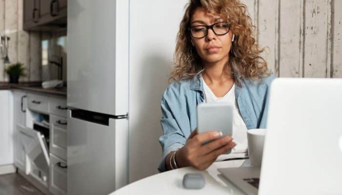 Woman looking at a smartphone while sitting beside a laptop at home.