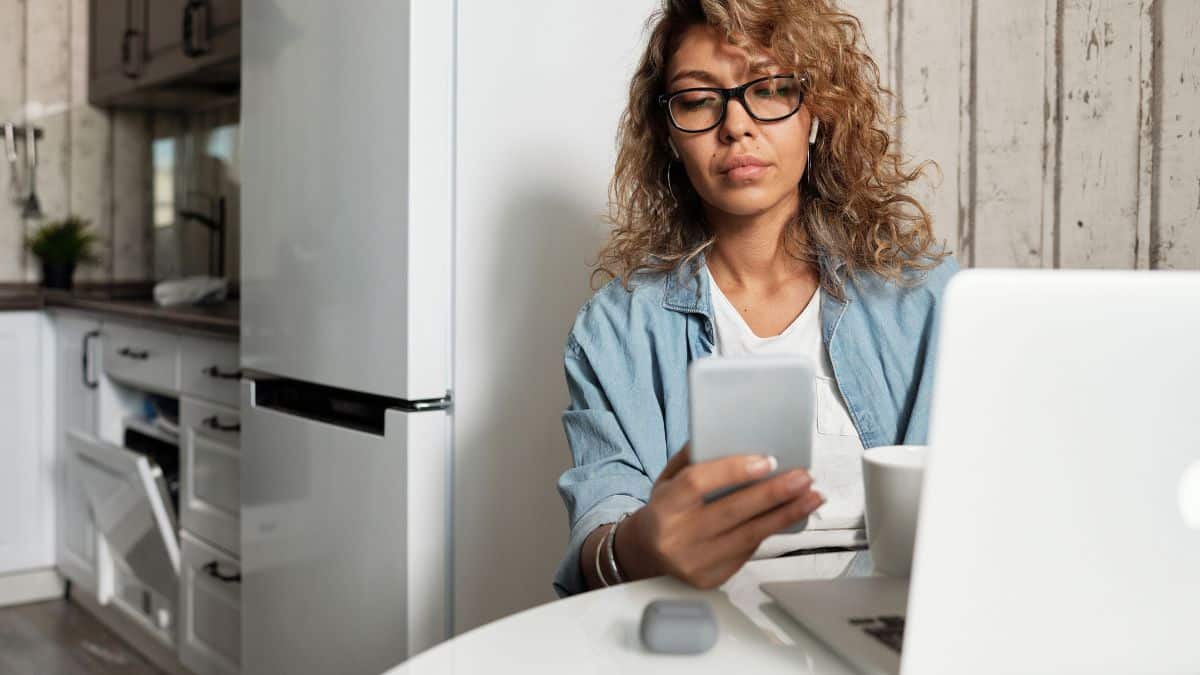 Woman looking at a smartphone while sitting beside a laptop at home.